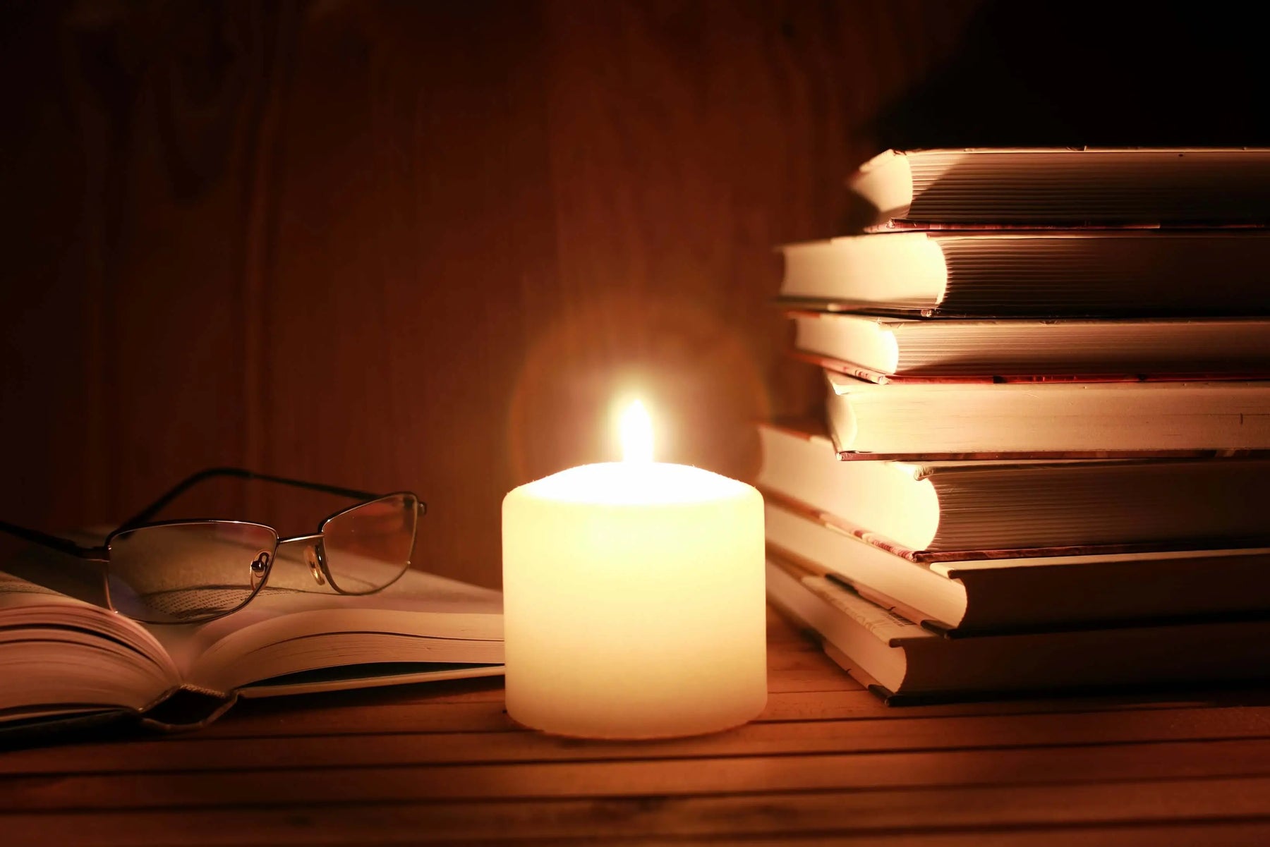 Candle illuminating stacked books and reading glasses on a wooden table.