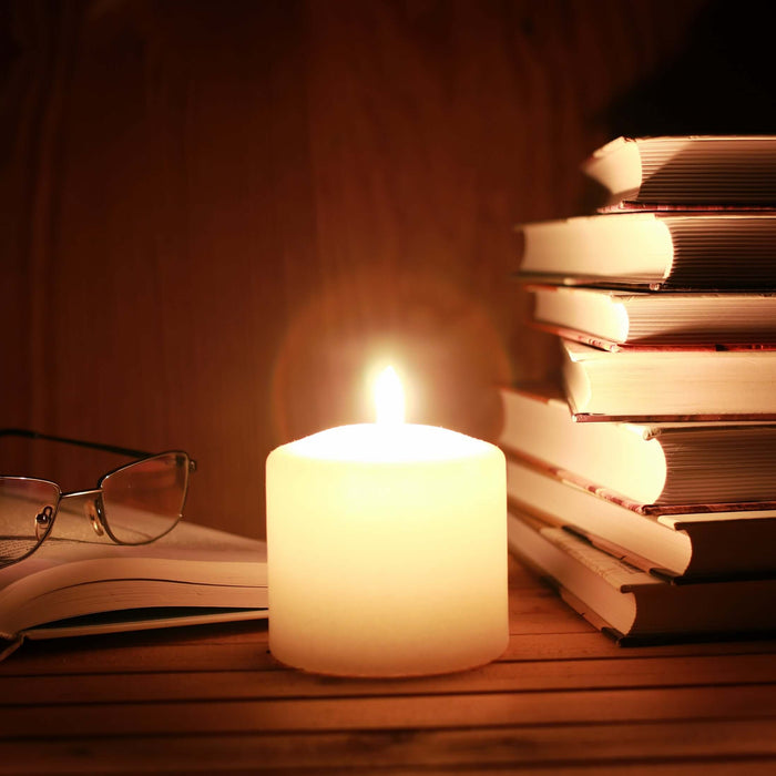 Candle illuminating stacked books and reading glasses on a wooden table.